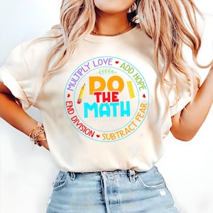May include: Cream-colored t-shirt with a colorful graphic that says "Do The Math" in a circle with the words "Multiply Love, Add Hope, Subtract Fear, End Division". A pencil and ruler are also in the design.