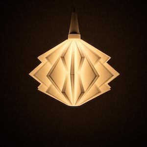 Könnte beinhalten: Eine moderne, geometrische Pendelleuchte. Der weiße Schirm hat ein geschichtetes, rautenförmiges Design und strahlt ein warmes, einladendes Licht aus. Die Leuchte hängt an einem dünnen Kabel vor einem dunklen Hintergrund und erzeugt einen starken Kontrast.