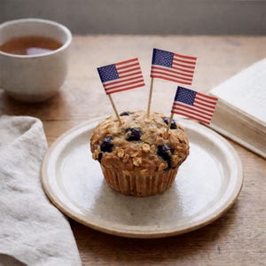May include: A blueberry muffin topped with three miniature American flags on a white plate. A cup of tea and an open book are in the background, all set on a wooden table. The muffin is a baked good.