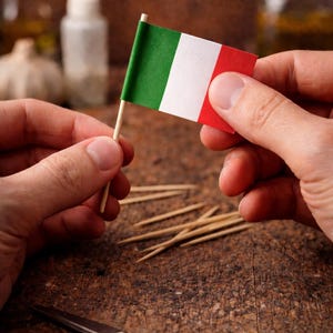 Könnte beinhalten: Eine kleine italienische Flagge mit grünen, weißen und roten Streifen wird zwischen zwei Händen gehalten. Die Flagge ist an einem kleinen Holzstab befestigt. Eine Schere und mehrere Zahnstocher liegen auf einer Holzoberfläche.