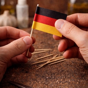 Könnte beinhalten: Eine kleine deutsche Flagge mit schwarzen, roten und gelben Streifen wird gehalten. Die Flagge ist an einem Holz-Zahnstocher befestigt. Verstreute Zahnstocher und eine Schere liegen auf einer Holzoberfläche.