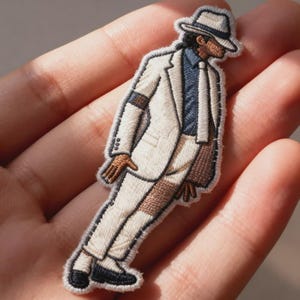 May include: An embroidered patch depicting a figure wearing a white suit, a dark blue shirt, a white tie, and a white fedora hat with a black band. The figure is shown in a leaning pose, wearing black shoes and a black armband on the right sleeve.
