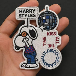 May include: An embroidered patch featuring a white cartoon dog wearing orange glasses, a red shirt with blue dots, and blue jeans. Above the dog is a sign reading HARRY STYLES and a disco ball. Text reads KISS ALL THE TIME OCCASIONALLY DISCO.