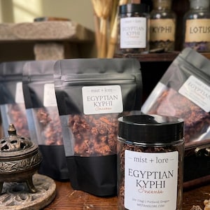 May include: An assortment of Egyptian Kyphi incense products. The image features clear glass jars and black pouches filled with dried incense. The labels read "Egyptian Kyphi Incense" and "mist + lore". A decorative incense burner is also visible.