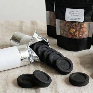 May include: Incense supplies are displayed on a neutral surface. Several rolls of charcoal are wrapped in silver foil with a white label. Black incense discs are visible, along with bags of incense and a bowl with a wooden spoon.