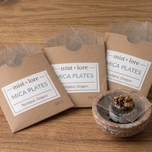 May include: Three brown paper envelopes containing transparent mica plates, each labeled with the text mist + lore MICA PLATES Portland, Oregon. One clear square plate rests on top of a small, round, mottled stone bowl containing dark material.