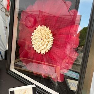 Puede incluir: Una obra de arte enmarcada que presenta una gran flor de plumas rojas vibrantes con un centro de color crema. La obra de arte se exhibe en un marco negro con un passe-partout blanco. El tarjetero en primer plano tiene el texto "La Plume Amienoise".