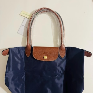 Puede incluir: Bolso tote azul marino con detalles en cuero marrón. El bolso tiene una solapa de cuero marrón con cierre de botón dorado. Las asas son de cuero marrón. Una etiqueta con el nombre de la marca está adjunta.