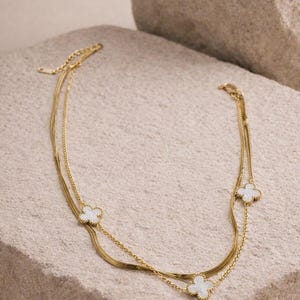 K&ouml;nnte beinhalten: Eine goldfarbene Halskette mit drei Str&auml;ngen, die jeweils mit einem wei&szlig;en, kleeblattf&ouml;rmigen Anh&auml;nger versehen sind. Die Halskette wird auf einer strukturierten, hellfarbigen Steinoberfl&auml;che pr&auml;sentiert, die ihr zartes Design und ihre elegante Ausstrahlung hervorhebt.