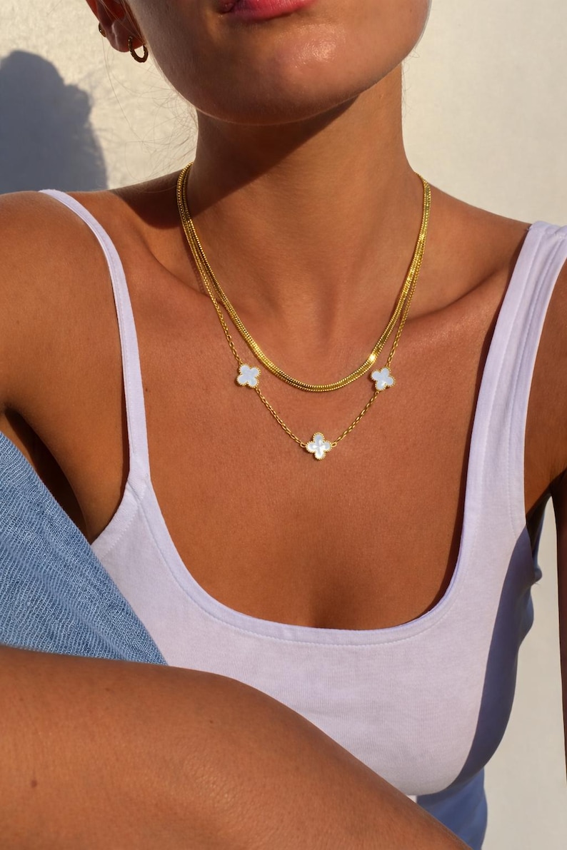 K&ouml;nnte beinhalten: Zwei goldene Halsketten mit Kleeblatt-Anh&auml;ngern. Die obere Halskette ist eine einfache Kette, w&auml;hrend die untere Halskette eine zarte Kette mit drei wei&szlig;en Kleeblatt-Anh&auml;ngern aufweist. Die Halsketten sind &uuml;bereinander getragen.