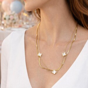 K&ouml;nnte beinhalten: Eine goldfarbene Halskette mit drei wei&szlig;en Kleeblatt-Anh&auml;ngern. Die Halskette hat zwei feine Kettenglieder und wird &uuml;ber einem wei&szlig;en Oberteil getragen. Der Hintergrund ist verschwommen, was auf eine festliche Veranstaltung hindeutet.