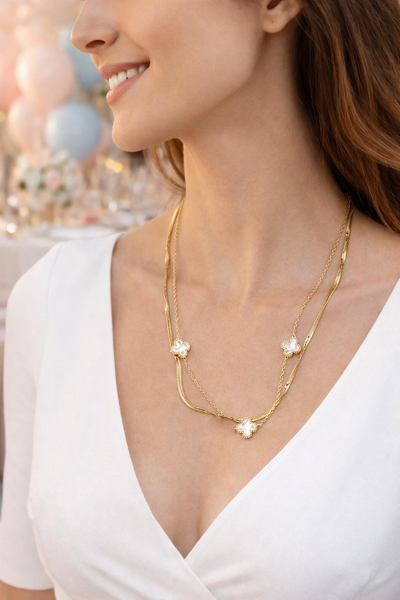 K&ouml;nnte beinhalten: Eine goldfarbene Halskette mit drei wei&szlig;en Kleeblatt-Anh&auml;ngern. Die Halskette hat zwei feine Kettenglieder und wird &uuml;ber einem wei&szlig;en Oberteil getragen. Der Hintergrund ist verschwommen, was auf eine festliche Veranstaltung hindeutet.