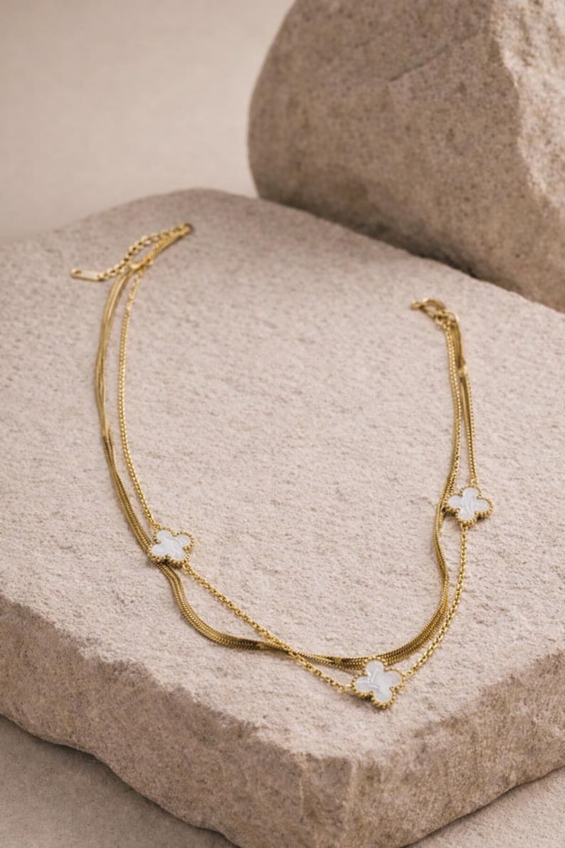 K&ouml;nnte beinhalten: Eine goldfarbene Halskette mit drei Str&auml;ngen, die jeweils mit einem wei&szlig;en, kleeblattf&ouml;rmigen Anh&auml;nger versehen sind. Die Halskette wird auf einer strukturierten, hellfarbigen Steinoberfl&auml;che pr&auml;sentiert, die ihr zartes Design und ihre elegante Ausstrahlung hervorhebt.