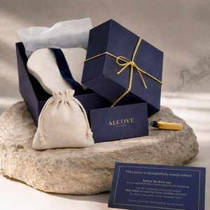 K&ouml;nnte beinhalten: Marineblaue Geschenkbox mit goldener Schleife, ge&ouml;ffnet, um einen wei&szlig;en Gegenstand und einen kleinen beigefarbenen Kordelzugbeutel zu enth&uuml;llen. Eine Visitenkarte mit dem Wort "ALCOVE" ist sichtbar. Ein brennender Holzstab liegt auf einem Stein.