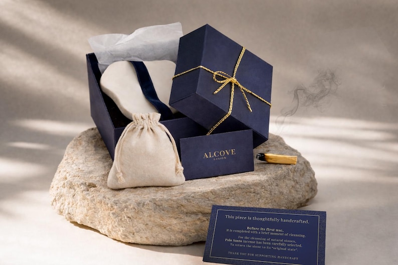 K&ouml;nnte beinhalten: Marineblaue Geschenkbox mit goldener Schleife, ge&ouml;ffnet, um einen wei&szlig;en Gegenstand und einen kleinen beigefarbenen Kordelzugbeutel zu enth&uuml;llen. Eine Visitenkarte mit dem Wort "ALCOVE" ist sichtbar. Ein brennender Holzstab liegt auf einem Stein.