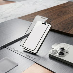 Op de afbeelding: Een doorzichtige glazen schermbeschermer met een zwarte rand en een ronde beschermer voor de cameralens. De verpakking is gelabeld met ULTRA-PROTECTION KIT. De items liggen naast een smartphone en een klein grijs reinigingsdoekje.
