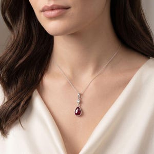 Op de afbeelding: Een zilveren ketting met een druppelvormige, dieprode edelsteen hanger aan een fijne ketting. Boven de druppelvormige steen bevindt zich een kleine, ronde, helder gefacetteerde steen in een zilveren zetting die de hanger verbindt.