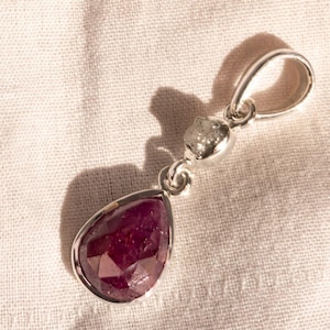 Peut inclure: Un pendentif en argent avec une pierre précieuse rubis rouge foncé en forme de poire à facettes, suspendue sous un petit diamant blanc brut. Le pendentif comprend une bélière en argent poli au sommet pour une chaîne ou un cordon.