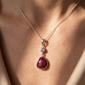 Op de afbeelding: Een zilveren hangerketting met een gefacetteerde, peervormige dieprode edelsteen die onder een kleine, ronde, helderwitte steen hangt. De hanger hangt aan een fijne zilveren ketting en heeft een gepolijste metalen zetting en een klassiek ontwerp.