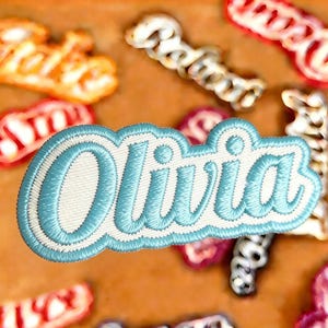 Könnte beinhalten: Gesticktes Namensschild mit dem Namen "Olivia" in hellblauem Faden und weißem Rand. Weitere Namensschilder in verschiedenen Farben sind sichtbar. Diese Aufnäher sind wahrscheinlich für Kleidung oder Accessoires bestimmt.