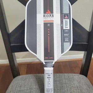 May include: A Joola Pro Perseus pickleball paddle with a black and white design. The paddle has the text "JOOLA PRO PERSEUS" and "16 MM" printed on it. The handle has the text "REMOVE FOIL TAP HERE".