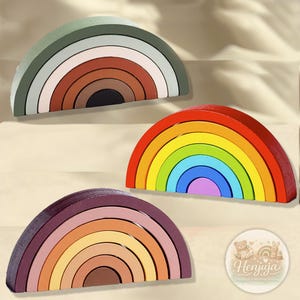 Könnte beinhalten: Drei Holz-Regenbogen-Stapler in Erdtönen und leuchtenden Farben. Die Stapler sind in Grün-, Braun-, Rot-, Orange-, Gelb-, Blau- und Lilatönen gehalten. Jeder Regenbogen hat mehrere Schichten und ist ein lehrreiches Spielzeug.