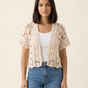 May include: A cream-colored, open-front crochet cardigan with short sleeves. The cardigan features an intricate openwork design and is worn over a white tank top and blue jeans. The overall style is casual and handmade.