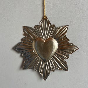 Puede incluir: Un adorno metálico plateado en forma de corazón con un diseño de estrella. El corazón está en el centro, rodeado de líneas radiales. Se adjunta una cuerda dorada para colgarlo. El adorno se muestra contra una pared gris claro lisa.