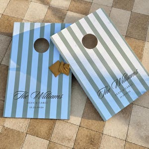 Könnte beinhalten: Zwei rechteckige Cornhole-Boards mit vertikalen hellblauen und weißen Streifen. Jedes Board hat ein kreisförmiges Loch oben und ist mit dem Text The Williams, Sean & Carli sowie 10.23.0026 personalisiert. Zwei senfgelbe Wurfsäckchen liegen auf einem Board.