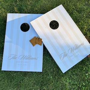 May include: Two rectangular cornhole boards featuring a light blue and white vertical striped pattern. Each board has a circular hole and is personalised with the text The Williams, Sean & Carli, and 10.24.2026. Two mustard yellow bean bags are included.