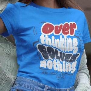 Puede incluir: Una camiseta azul con el texto "Overthinking Solves Nothing" en un diseño blanco, rojo y azul marino. La camiseta también presenta el texto "Est. 2026" y un pequeño gráfico.