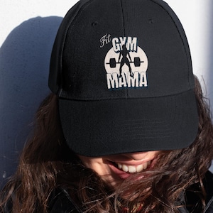 Puede incluir: Gorra de béisbol negra con el texto "Fit Gym Mama" en blanco y azul. El diseño presenta una silueta de levantador de pesas dentro de un círculo blanco. La gorra está hecha de tela duradera y tiene una visera curva.