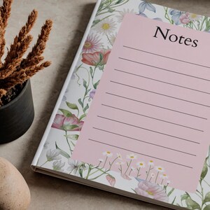 Può includere: Un taccuino a tema floreale con una pagina di note rosa. La copertina presenta uno sfondo bianco con fiori ad acquerello rosa, viola e verdi. La pagina delle note rosa ha la parola "Note" e righe.
