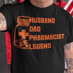 May include: Black t-shirt with orange text and graphics. The text reads "HUSBAND DAD PHARMACIST LEGEND" with an illustration of a pill bottle and pills. The shirt is short-sleeved.