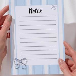 Può includere: Un blocco note con sfondo a righe azzurre e bianche. Il blocco note ha una sezione rettangolare bianca con linee orizzontali per la scrittura. La parola "Notes" è scritta in corsivo nero in alto, con un'illustrazione di fiocco. Un'illustrazione di fiocco è anche in basso.