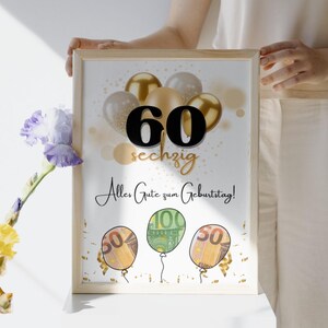 May include: A framed print with a birthday theme. The print features the number 60 in black, with the word "sechzig" below it in gold. Gold and silver balloons are in the background. The text "Alles Gute zum Geburtstag!" is written in script. Balloons with money are at the bottom.