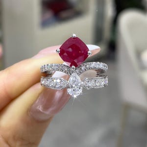 May include: A two-piece ring set featuring a large, square-cut ruby gemstone atop a pear-shaped diamond. The rings are set in a silver band, with smaller diamonds accenting the bands. The ring is held up by a person's hand.