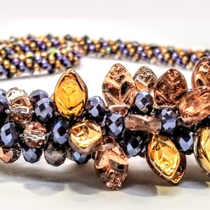 Può includere: Una collana di perline con un designo floreale. La collana è realizzata con perline blu scuro, argento e oro. I fiori sono realizzati con perline di rame e oro.