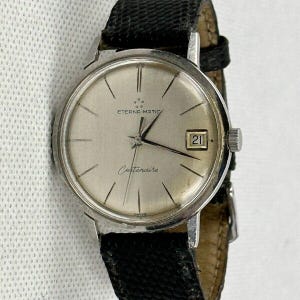 May include: A vintage Eterna-Matic Centenaire wristwatch with a silver dial and black leather band. The watch face displays the date and the words "Eterna-Matic" and "Centenaire". The watch has a silver-colored case and a textured black leather band.