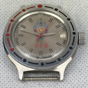 Puede incluir: Reloj de pulsera plateado con esfera gris que presenta un emblema de estrella roja, escudo y espada. El reloj tiene una ventana de fecha y las letras cirílicas "КГБ" en rojo. El bisel tiene detalles en rojo y negro.