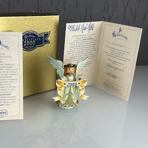 Puede incluir: Una figura de ángel coleccionable de la serie "Good Dreams", sosteniendo una pancarta con "2000". El ángel tiene alas y viste un vestido dorado y blanco. La figura se exhibe con su caja y certificado originales.