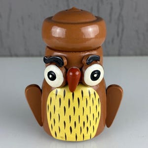 May include: A brown wooden owl figurine with large white eyes, a red beak, and black eyebrows. The owl has a yellow chest with black vertical lines and small brown wings. The top of the owl's head is a separate piece.