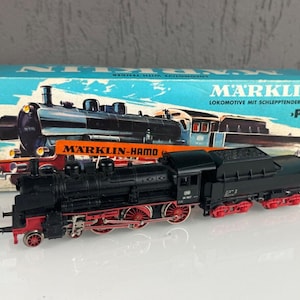 Puede incluir: Un modelo de tren negro con ruedas y detalles rojos, y un ténder de carbón. El tren está etiquetado como "MÄRKLIN-HAMO" y se encuentra sobre una caja azul y blanca con ilustraciones de trenes.