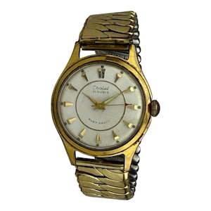 Könnte beinhalten: Vintage goldfarbene Chalet Armbanduhr mit weißem Zifferblatt. Die Uhr hat goldfarbene Stundenmarkierungen, Zeiger und einen Sekundenzeiger. Das Armband ist aus goldfarbenem, flexiblem Metall. Auf dem Zifferblatt steht "Chalet 21 Rubis" und "Ruby Shock".