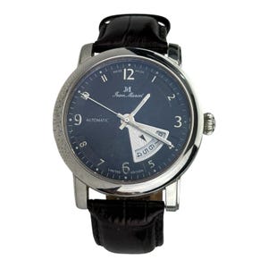 May include: A silver-toned Jean Marcel automatic wristwatch with a dark blue face and black leather band. The watch face features the text "SWISS MADE", "Jean Marcel", and "AUTOMATIC". The watch has a date display and a textured bezel.