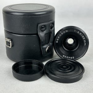 Può includere: Un obiettivo per fotocamera Berogon nero 1:3,5/35 mm con un copriobiettivo abbinato e una custodia in pelle nera. L'obiettivo ha il numero di serie 294226. La custodia è cilindrica con coperchio incernierato e maniglia.