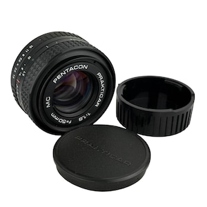 Pode incluir: Uma lente de câmera Pentacon Prakticar preta de 50 mm f/1.8 com uma tampa de lente e um anel de proteção. A lente tem texto branco que diz "Pentacon Prakticar" e "f=50mm 1:1.8". A tampa da lente também tem o texto "Prakticar".