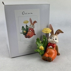 Op de afbeelding: Een keramieken paashaasfiguur met een paardenbloem. Het konijn draagt een oranje overall en een rode vlinderdas. Het beeldje staat naast een witte doos met het woord "Ostern" en een afbeelding van het konijn.
