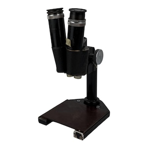 May include: A vintage black microscope with two eyepieces and a dark brown base. The microscope has a vertical support and a focusing knob. The base is rectangular with a slightly angled front edge. The eyepieces have silver rings.