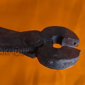 May include: Close-up of a dark, rusty metal tool with curved jaws and long handles. The tool, likely tongs or pliers, is set against an orange background. The text "THE KNIGHT ARMOURY" is visible on the handle.